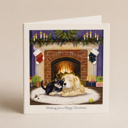 Square Christmas card featuring a cat and dog cuddling on a rug before a glowing fireplace with festive stockings.