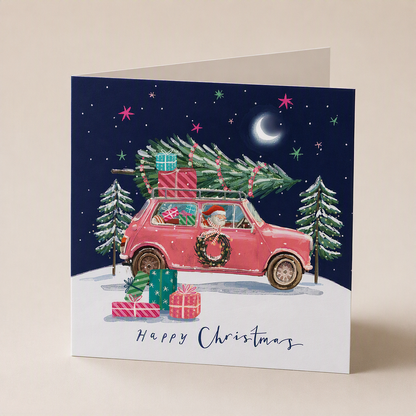 Square Christmas card featuring a pink car with gifts and a tree in a snowy landscape under a starry night sky.