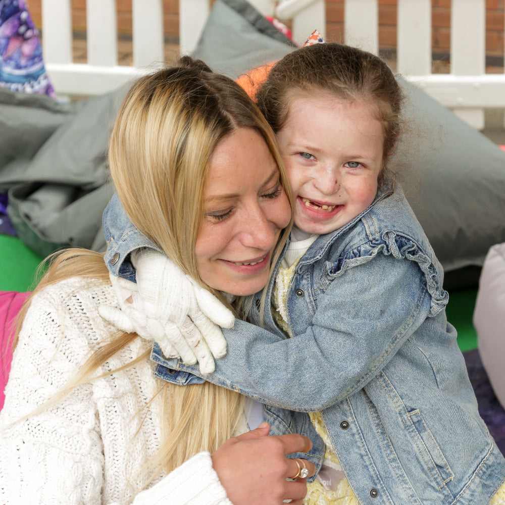 A woman and a young girl hugging in a cozy lounge setting with colorful cushions, pillows, and a white fence in the background.