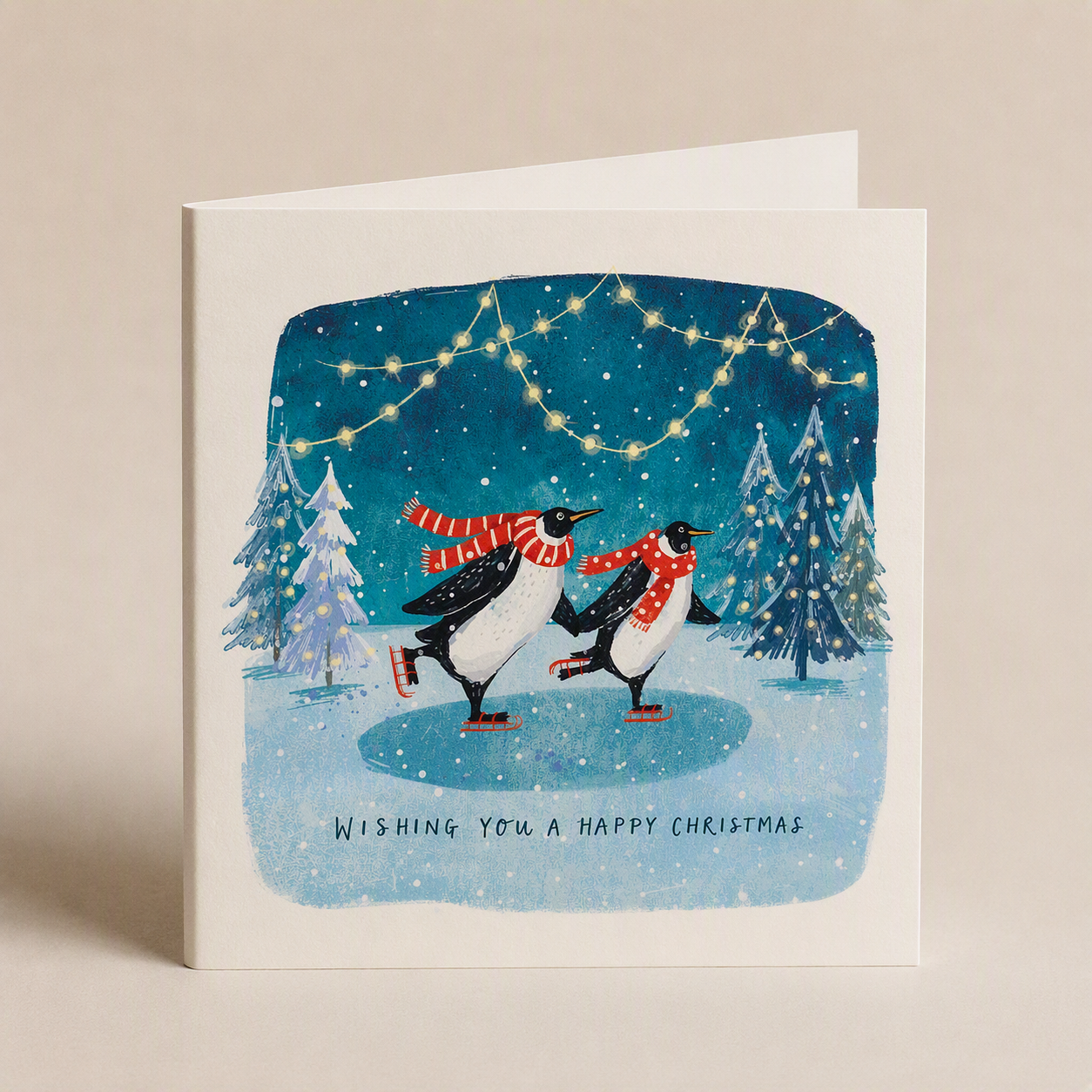 Two penguins in red scarves ice skating on a snowy pond with fairy lights on a square satin-finish Christmas card.